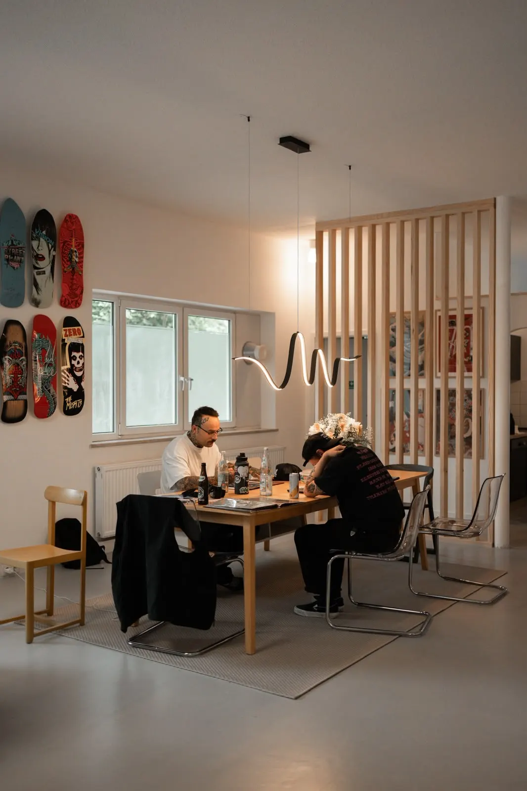 Zeichentisch unter der Lampe, dahinter die Holzlamellen-Trennwand und Skateboards